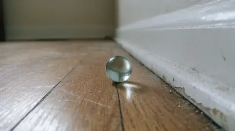 Glass marble that rolled toward the baseboard on a wood floor, with a gap under the baseboard